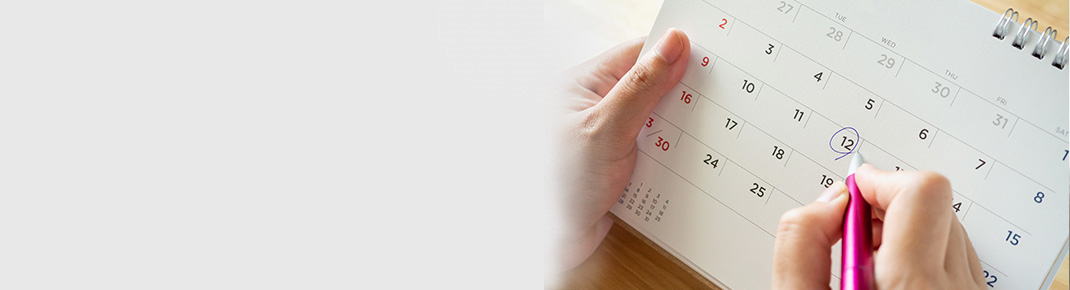 Mulher de óculos, vestindo uma camisa azul escura, se inclina sobre uma cama branca com um sorriso, parecendo pensativa. / Close-up de um calendário com a mão de uma mulher marcando um dia com uma caneta